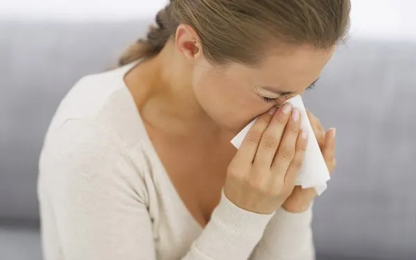 A woman sneezing into a tissue while seated on a sofa, appearing to have a cold or allergies.
