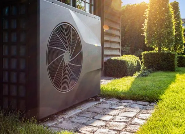 Outdoor heat pump unit beside a house on a cobblestone path in a sunny, landscaped yard with trimmed hedges