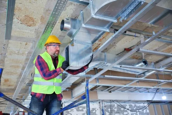 A construction worker on scaffolding installs metal HVAC ductwork in a building ceiling, wearing hard hat and high-vis vest.