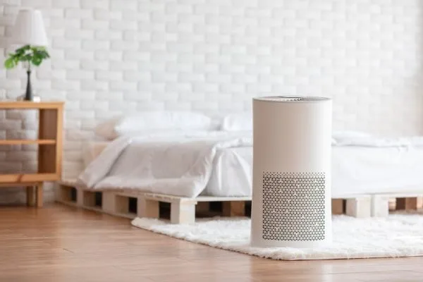 White cylindrical air purifier on rug in a minimalist bedroom with low pallet bed and wooden floors