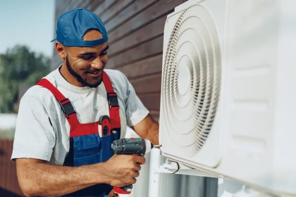 A man HVAC technician in a cap and overalls installing an outdoor air conditioning unit with a cordless drill.