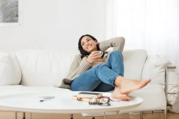 A woman relaxing barefoot on a white couch, smiling and holding a mug, with books and glasses on a coffee table.