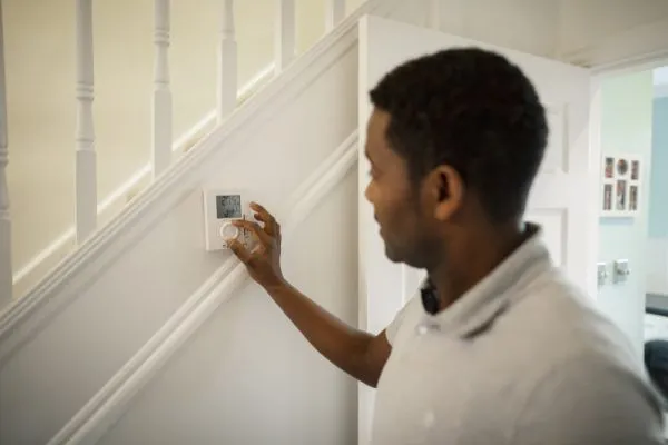 A man adjusts a smart thermostat mounted on a hallway wall next to a staircase in a modern home.