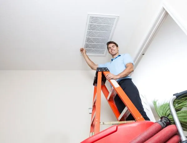 A man on a ladder inspecting a ceiling air vent during HVAC maintenance in a bright room