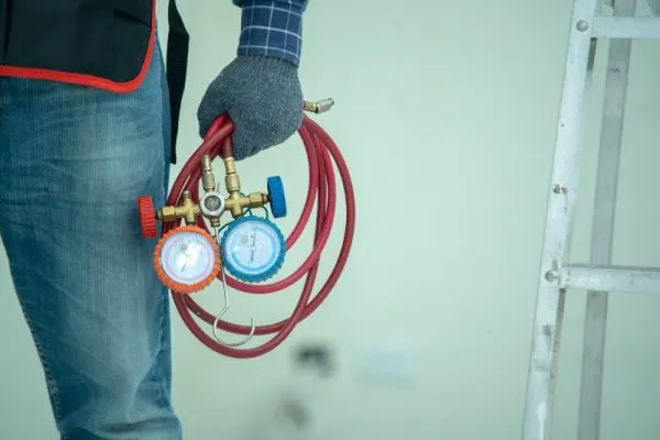 A person holding an HVAC manifold gauge set with red hoses beside a ladder, wearing gloves and jeans